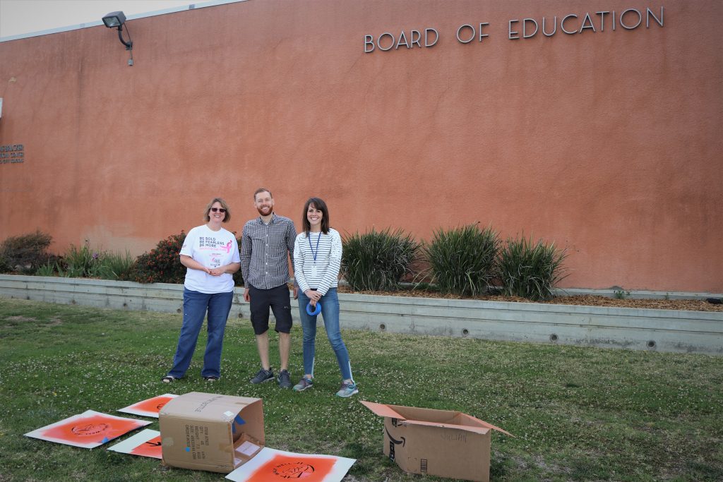 three people standing on grass in front of the board of education